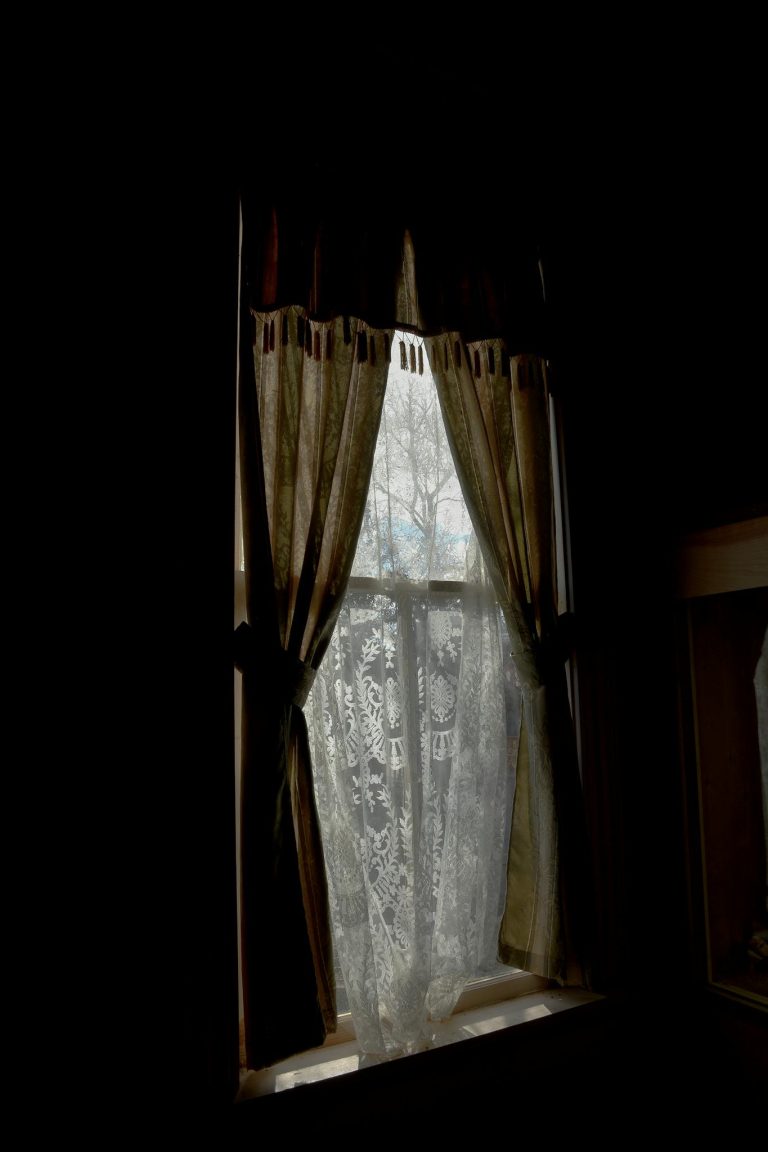 Warm light filters through lace and draped curtains, creating a cozy interior scene.